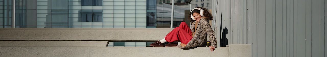 Person sitting on a concrete bench in a modern urban setting, wearing white earmuffs, a checked oversized coat, red trousers, and brown slip-on shoes.