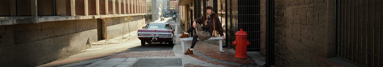 Person sitting on a concrete bench in a modern urban setting, wearing white earmuffs, a checked oversized coat, red trousers, and brown slip-on shoes. Large white text on the left reads: 'THE NEW COZY'.