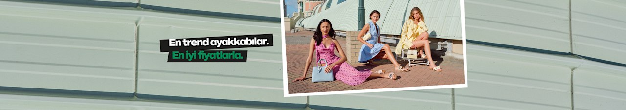 Three people pose outdoors on a paved area in front of a light green, corrugated wall. One person sits on the ground wearing a pink outfit and holding a small blue handbag, while two others sit on folding chairs wearing light summer outfits in blue and yellow. All three are wearing sandals. A text overlay on the left reads: “The most beautiful details. The best prices.