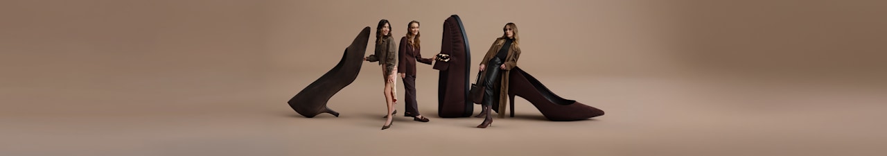 Fashion campaign image on a beige background featuring oversized chocolate brown loafers and high heels. Three models are posed around the shoes, wearing coordinated outfits in brown, black, and neutral tones. The footwear and styling create a sophisticated autumn fashion scene with a focus on rich brown shades.