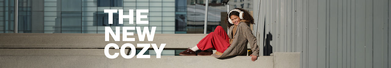 Person sitting on a concrete bench in a modern urban setting, wearing white earmuffs, a checked oversized coat, red trousers, and brown slip-on shoes. Large white text on the left reads: 'THE NEW COZY'.
