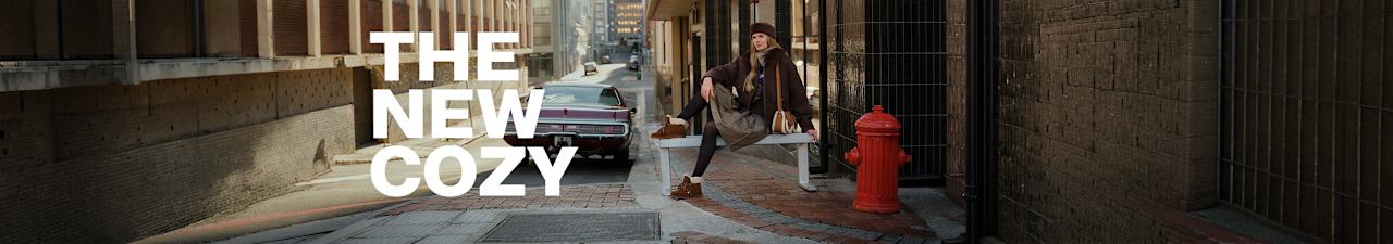 Person sitting on a white bench at a city street corner, wearing a dark jacket, knit hat, skirt, tights, and brown shoes. Large white text on the left reads: 'THE NEW COZY'. A red fire hydrant and a vintage car are visible in the background.