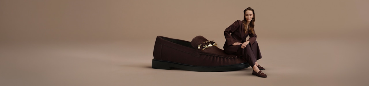 A campaign image on a neutral beige background features an oversized dark brown loafer with a decorative gold-tone chain detail. A person wearing a coordinated dark brown outfit and matching loafers is seated beside the shoe. The studio composition highlights the footwear's classic silhouette, rich brown color, and metallic chain accent.