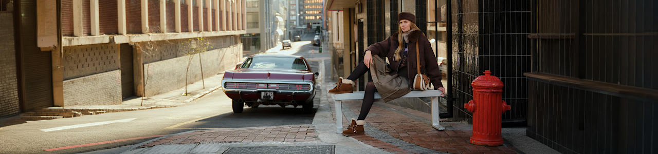 Person sitting on a white bench along a city street, wearing a brown jacket, knit hat, skirt, tights, and brown shoes. A red fire hydrant and a vintage car are visible nearby.