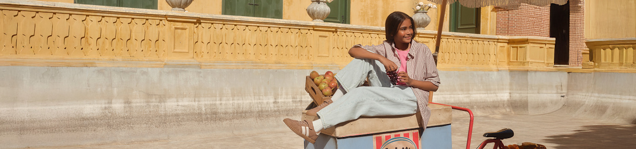 A child sitting on an ice-cream cart in a sunny courtyard, wearing light pants and tan sneakers with a strap.
