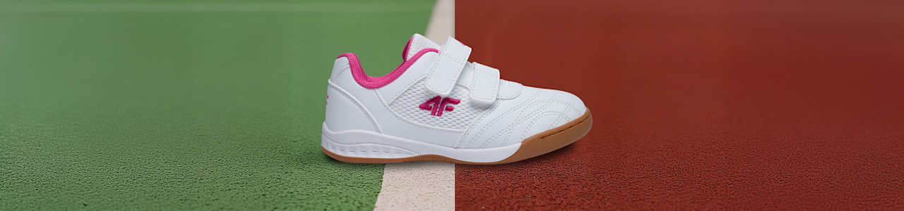 A white children’s sneaker with pink accents and two Velcro straps is placed on a sports court, positioned over a white line that separates the green left side from the red right side.