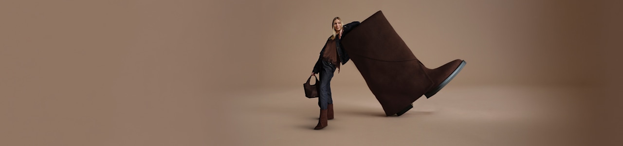 A campaign image on a neutral beige background features a person standing beside an oversized dark brown ankle boot. The person is styled in a dark denim outfit with matching brown ankle boots and carries a dark brown handbag. The enlarged boot dominates the composition, creating a fashion-focused studio visual that highlights the footwear and coordinated accessories.