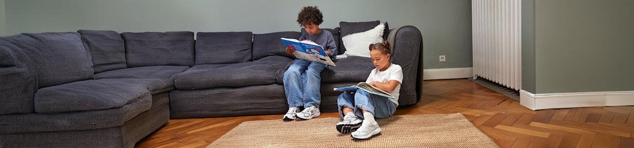 Two children sit on a woven rug in a cozy living room, each looking through a magazine. The room features a dark sofa, a wooden parquet floor, and a neutral-colored rug. Both children wear casual outfits and New Balance sneakers. The scene highlights a relaxed indoor lifestyle setting focused on everyday comfort and kids’ footwear.