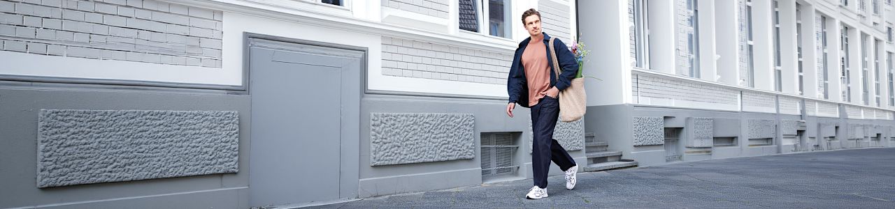 A person walks along a city sidewalk in front of a light-colored residential building, carrying a canvas tote bag. The outfit consists of a coral-colored top, a dark jacket, dark trousers, and white New Balance sneakers. The urban setting and casual styling create a relaxed, everyday lifestyle look.
