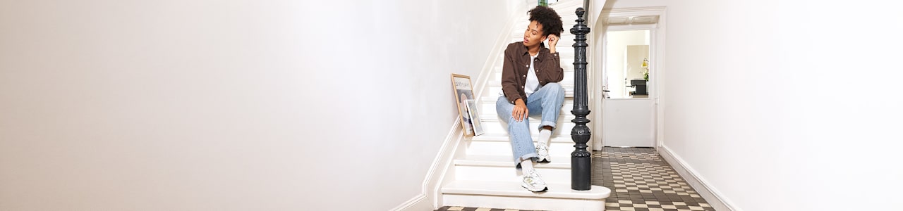 A person sits on a white staircase in a bright home interior, wearing a brown overshirt, light blue jeans, and white New Balance sneakers. Framed artwork is displayed along the staircase, while a black banister and patterned floor tiles add contrast to the light surroundings. The image highlights a relaxed lifestyle look and casual footwear styling.