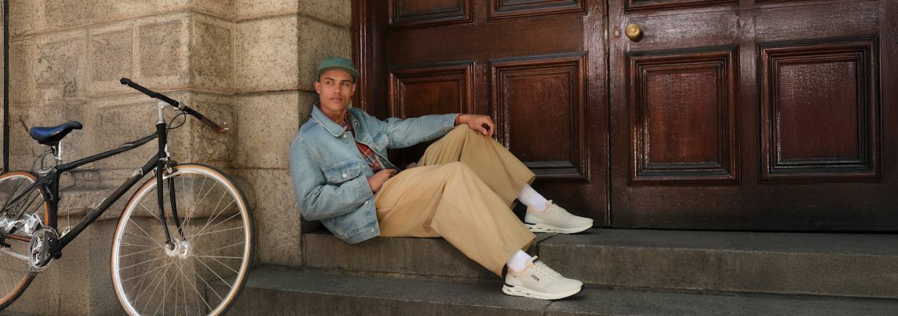 A person in a denim jacket, plaid shirt, wide trousers, and light sneakers beside a bicycle in an urban stone-arched setting.