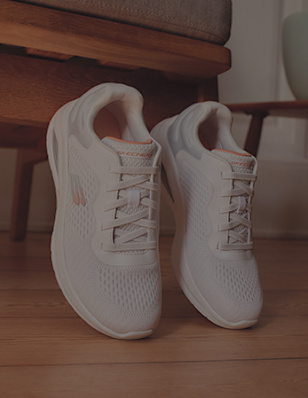 White Skechers sneakers with light pastel accents positioned upright on a wooden floor beneath a cushioned chair.