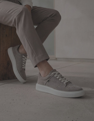 Person seated indoors wearing light grey suede low-top sneakers with white soles and beige laces, paired with taupe trousers, feet resting on a smooth concrete floor beside a wooden block