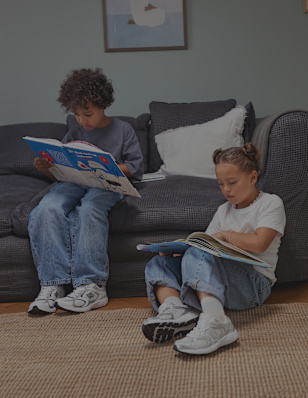 Two children sit on a woven rug in a cozy living room, each looking through a magazine. The room features a dark sofa, a wooden parquet floor, and a neutral-colored rug. Both children wear casual outfits and New Balance sneakers. The scene highlights a relaxed indoor lifestyle setting focused on everyday comfort and kids’ footwear.