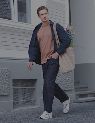 A person walks along a city sidewalk in front of a light-colored residential building, carrying a canvas tote bag. The outfit consists of a coral-colored top, a dark jacket, dark trousers, and white New Balance sneakers. The urban setting and casual styling create a relaxed, everyday lifestyle look.