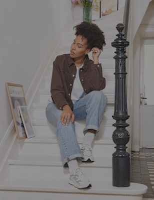 A person sits on a white staircase in a bright home interior, wearing a brown overshirt, light blue jeans, and white New Balance sneakers. Framed artwork is displayed along the staircase, while a black banister and patterned floor tiles add contrast to the light surroundings. The image highlights a relaxed lifestyle look and casual footwear styling.