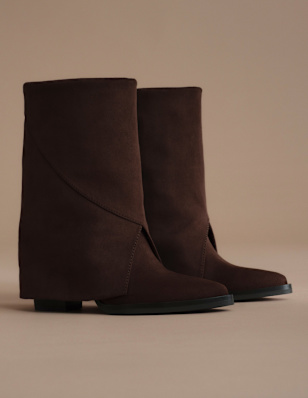 A pair of dark brown ankle boots is displayed against a neutral beige background. The boots feature a minimalist design with smooth suede-like material, a rounded toe, and low block heels. The clean studio setting highlights the rich brown color and understated silhouette of the footwear.