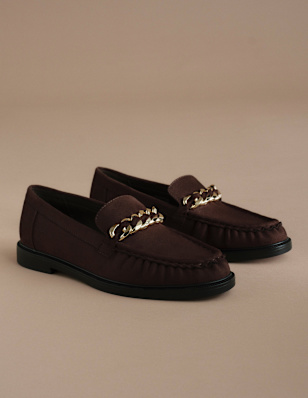 A pair of dark brown loafers is displayed on a neutral beige background. The shoes feature black soles, subtle stitched detailing along the upper, and a decorative gold-tone chain across the front. The minimalist studio setting highlights the rich brown color and classic loafer silhouette.