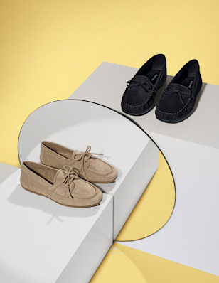 Two pairs of loafers displayed on white geometric platforms against a yellow background, featuring beige lace-up suede loafers in the foreground and black slip-on loafers positioned behind, styled in a minimalist studio setting.