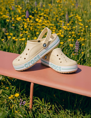 Pair of beige Crocs-style clogs placed on a bench outdoors in a sunny field.