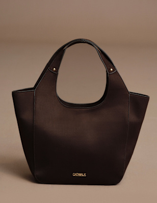 A dark brown handbag is displayed against a neutral beige background. The structured design features rounded top handles, a spacious silhouette, and subtle gold-tone branding on the front. The clean studio setting emphasizes the rich brown finish and minimalist shape.