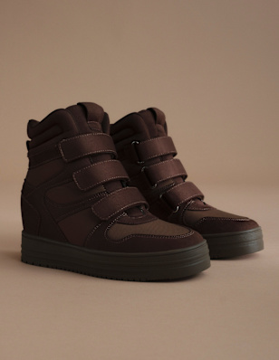A pair of dark brown high-top sneakers is displayed on a neutral beige background. The shoes feature a chunky sole, padded collar, and multiple hook-and-loop straps across the upper, combining a sporty look with autumn-inspired styling.