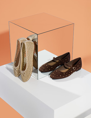 A pair of beige woven ballet flats and a pair of brown leather Mary Jane shoes with small gold studs are displayed on a white pedestal against a peach background, with a transparent cube placed between them.