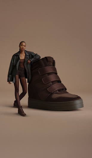 A campaign image on a neutral beige background features an oversized dark brown high-top sneaker with a chunky sole and multiple hook-and-loop straps. A person wearing a dark jacket, brown top, matching dark shorts, and coordinating high-top sneakers stands beside the shoe. The studio composition highlights the sneaker’s sporty silhouette and autumn-inspired color palette.