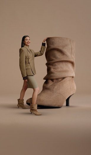 A campaign image on a neutral beige background features an oversized beige slouchy ankle boot with a soft suede-like texture. A person wearing a coordinated beige outfit and matching ankle boots stands beside the shoe with one hand resting on it. The composition emphasizes the boot’s gathered shaft, pointed toe, and slim heel.
