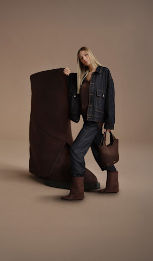 A campaign image on a neutral beige background features an oversized dark brown ankle boot. A person wearing a dark denim outfit, matching brown ankle boots, and a dark brown handbag stands beside the shoe. The fashion-focused studio composition highlights the coordinated footwear and accessories.