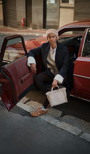Person sitting in the open doorway of a red vintage car, wearing a dark overcoat, light knit hat, layered knitwear, dark trousers, and brown lace-up shoes. A light-colored handbag is held in one hand.