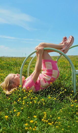 Child lying on a climbing frame wearing pink slide sandals in a meadow.