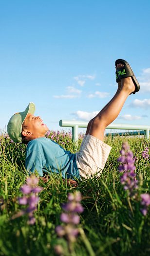 Child playing in grass wearing black slide sandals with green logo detail.