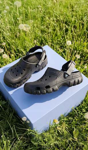 Pair of black clog-style sandals displayed on a pedestal in a green meadow.