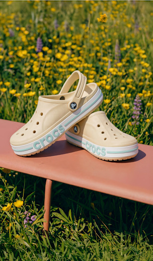 Pair of beige Crocs-style clogs placed on a bench outdoors in a sunny field.