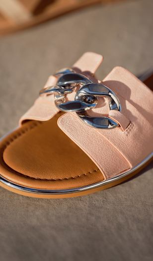 Close-up of a light pink slide sandal with a wide strap and a large metallic chain detail on top. The sandal has a soft, matte texture and a brown sole with a thin metallic edge. It is placed on a sunlit surface, highlighting the material and decorative feature.
