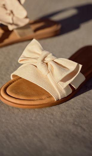 Close-up of a light beige slide sandal with a wide strap and a large bow detail on top. The sandal has a soft, textured surface and a brown sole. It is placed on a sunlit ground, highlighting the material and decorative design.