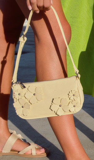 Close-up of a light yellow shoulder bag with a slim strap and floral appliqué details on the front. The bag is carried against a bright green skirt, with a sandal visible on the foot below. The scene is set outdoors in strong sunlight, highlighting the texture and decorative elements of the bag.