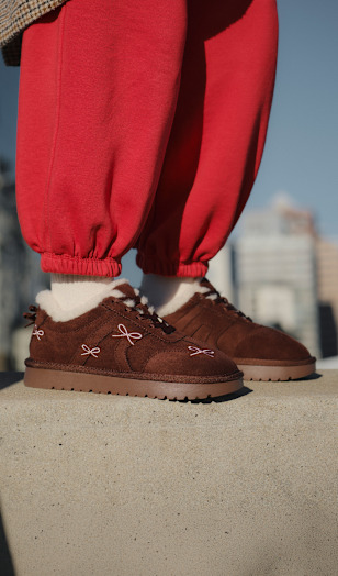 Close-up of brown suede slip-on shoes with white fleece lining, paired with white socks and bright red jogger-style trousers, standing on a concrete ledge.