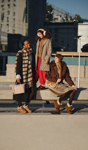 Three people gathered on a concrete bench in a modern urban setting, wearing layered cold-weather outfits and casual footwear.