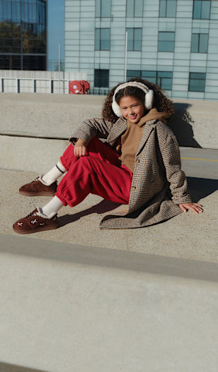 Person sitting on a concrete bench in a modern urban setting, wearing white earmuffs, a checked coat, a hoodie, red trousers, and brown slip-on shoes.