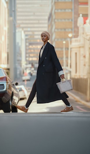 Person walking across a city street, wearing a long dark overcoat, light knit hat, dark trousers, and brown fleece-lined lace-up shoes, while carrying a light-colored handbag.