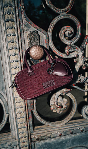Close-up of a burgundy FILA handbag with textured fabric, leather details, and a heart-shaped charm, displayed against an ornate wrought-iron gate. The compact bag is highlighted by dramatic lighting that emphasizes its rich color and vintage-inspired design.