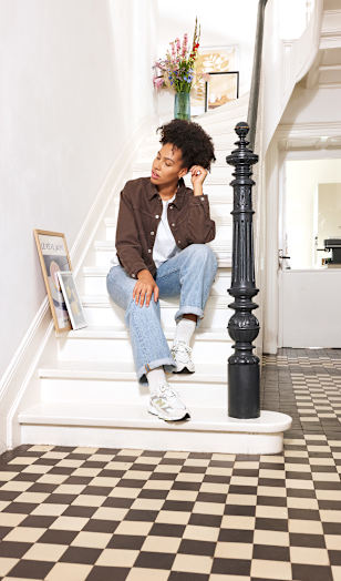 A person sits on a white staircase in a bright home interior, wearing a brown overshirt, light blue jeans, and white New Balance sneakers. Framed artwork is displayed along the staircase, while a black banister and patterned floor tiles add contrast to the light surroundings. The image highlights a relaxed lifestyle look and casual footwear styling.