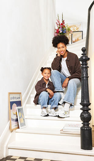 A person and a child sit together on a white staircase in a bright home interior. Both wear matching brown overshirts, light blue jeans, and white New Balance sneakers. Framed artwork leans against the wall beside the stairs, while colorful flowers and framed prints decorate the landing above. The scene highlights coordinated casual footwear and relaxed everyday style.