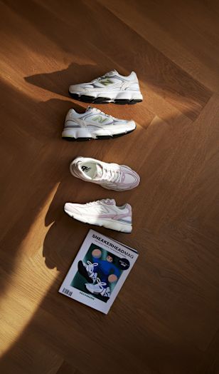Four New Balance sneakers in white with grey, black, and light pink accents are arranged on a warm-toned wooden floor. Natural sunlight casts soft shadows across the scene. The shoes are displayed in a spaced layout that highlights different colorways and design details. A copy of Sneakerheadmag is placed beside the sneakers, reinforcing the sporty lifestyle theme.