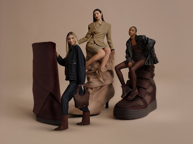 Three oversized brown and beige boots are displayed against a neutral beige background. Three fashion models pose around and on the shoes, wearing coordinated autumn-inspired outfits in shades of taupe, brown, and black. One model stands beside a slouchy beige heeled boot, another sits on a tall dark brown boot, and a third leans against a chunky brown strap boot. The image combines footwear and fashion styling in a clean studio setting.