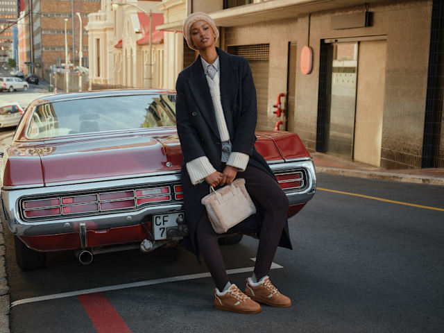 Person sitting on the rear of a red vintage car parked on a city street, wearing a long dark coat, knit hat, light-colored handbag, and casual shoes.