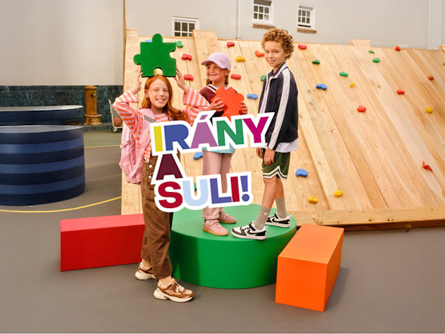Children posing with colorful school-themed letters in front of a climbing wall. Letters say "Irany a Suli".