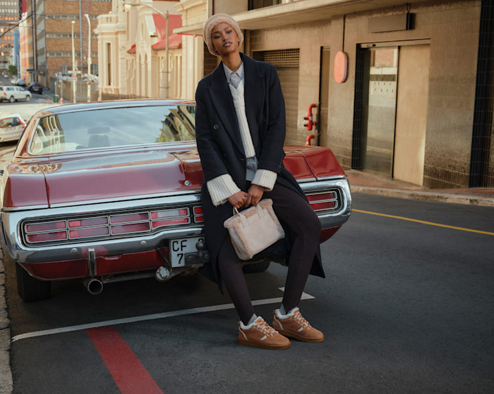 Person sitting on the rear of a red vintage car parked on a city street, wearing a long dark coat, knit hat, light-colored handbag, and casual shoes.